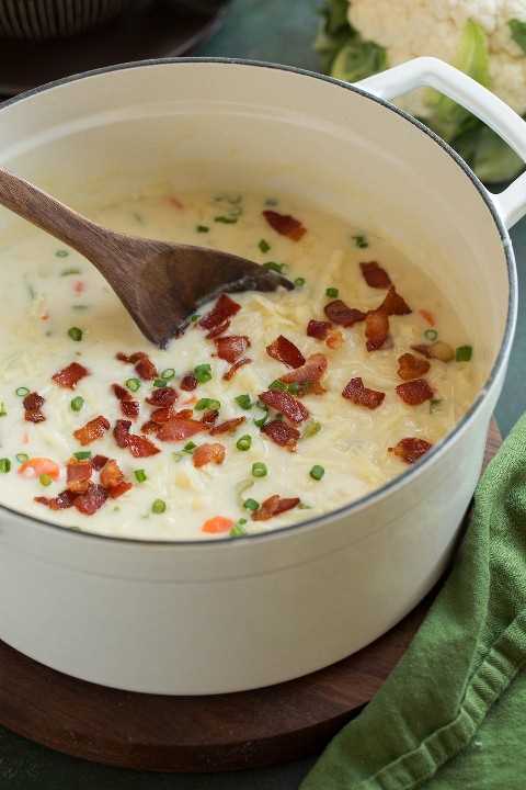 Olla llena de sopa de coliflor adornada con queso cheddar, tocino y cebolla verde.