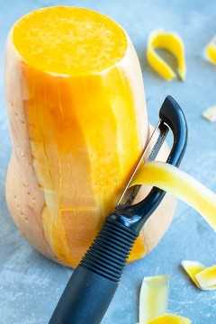 A potato peeler that is peeling the skin off of a large butternut winter squash.