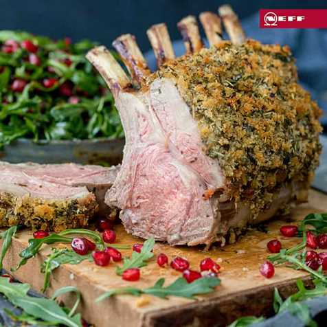 Herb crusted rack of lamb on a wooden board with a slice taken off - showing pink centre. Rocket and pomegranate seeds strewn around the board.