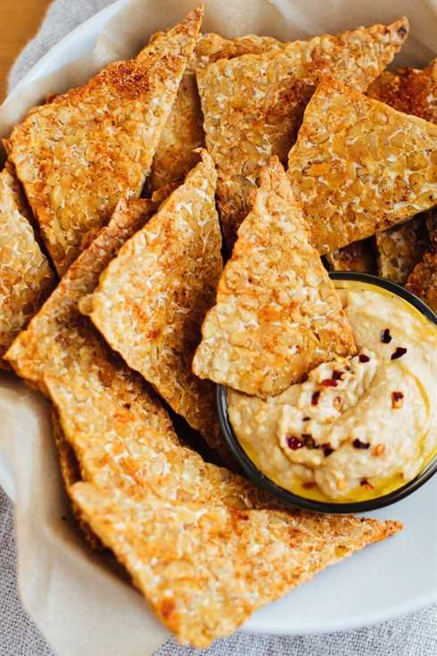 Close up of BBQ tempeh chips with hummus in a white bowl.
