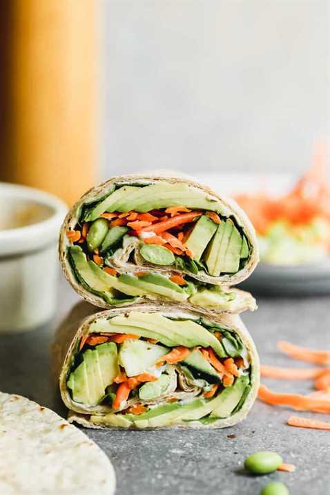 Two veggie wraps stacked on each other, filled with avocado, carrots, hummus and edamame.