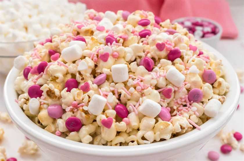 Close up of a bowl of Pretty Pink Party Popcorn sitting on a white surface surrounded by a bowl of mini marshmallows and a ramekin of Valentine