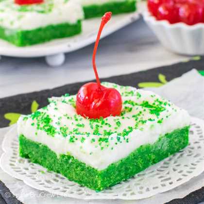 A white plate with a green shamrock shake sugar cookie bar topped with frosting, green sugar, and a cherry on it