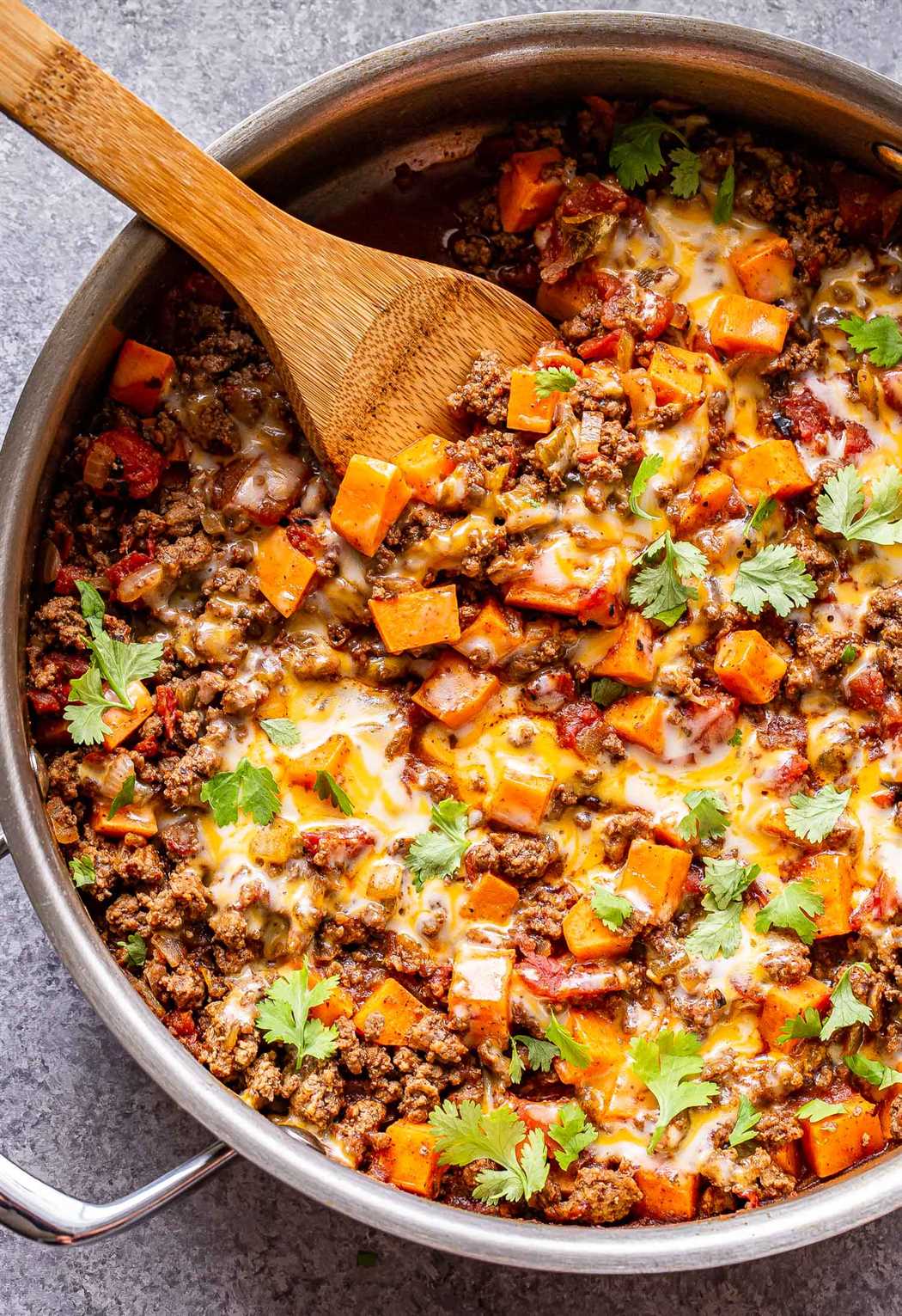 Overhead photo of a metal skillet filled with ground beef, tomatoes, sweet potatoes, southwest spices and topped with melted cheese and cilantro. Wooden serving spoon in the skillet.