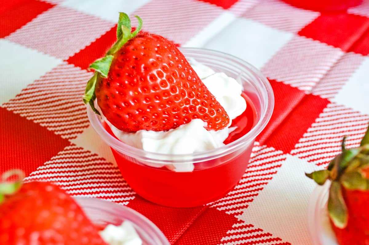 strawberry jello shot topped with whipped cream and a fresh strawberry, served in a plastic cup and on top of a red and white checkered tablecloth