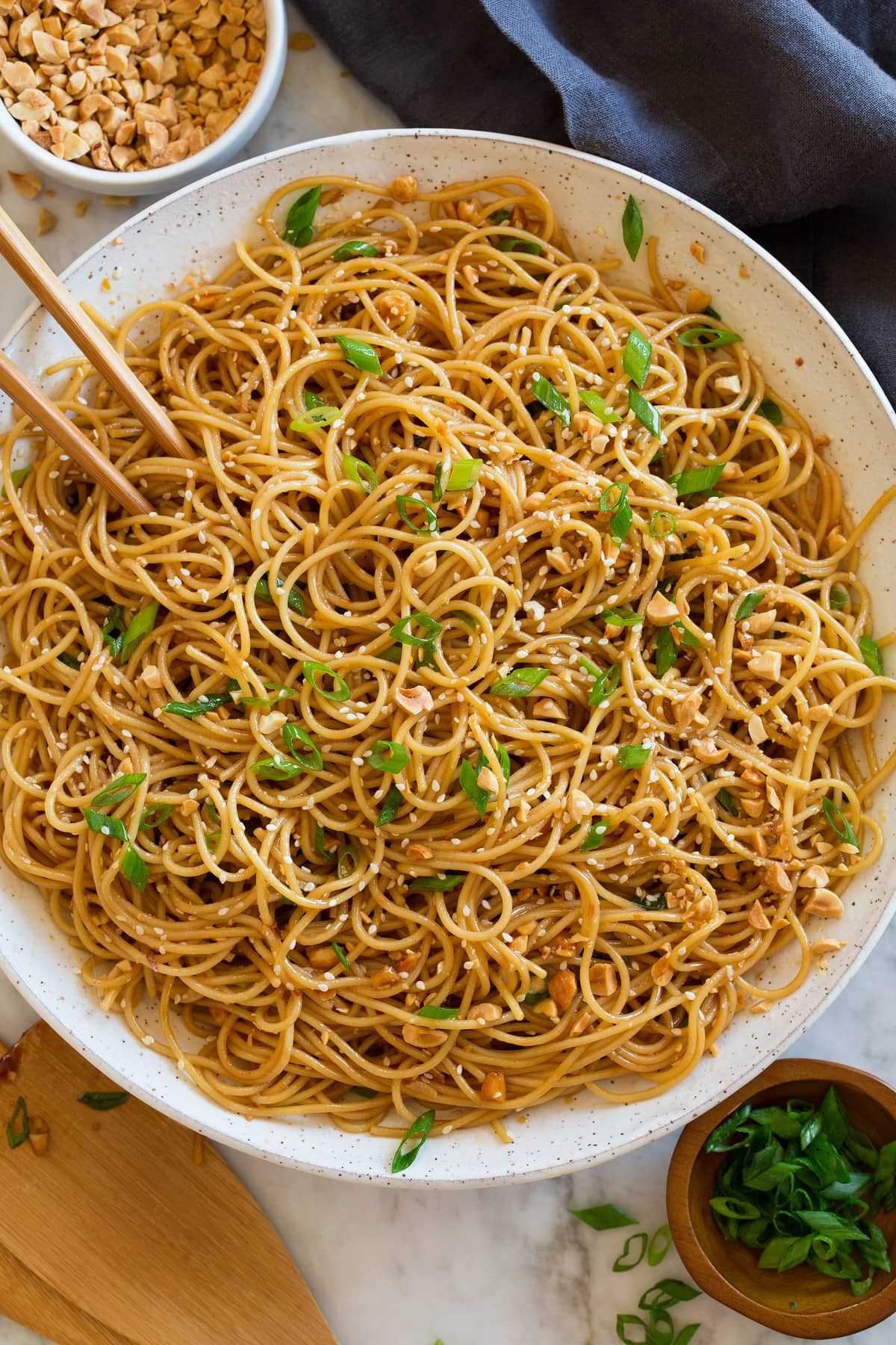 Photo: Bowl of sesame noodles topped with peanuts, sliced green onions and sesame seeds. Shown overhead in a white bowl with a dark napkin underneath and chopsticks and serving spoons to the side.