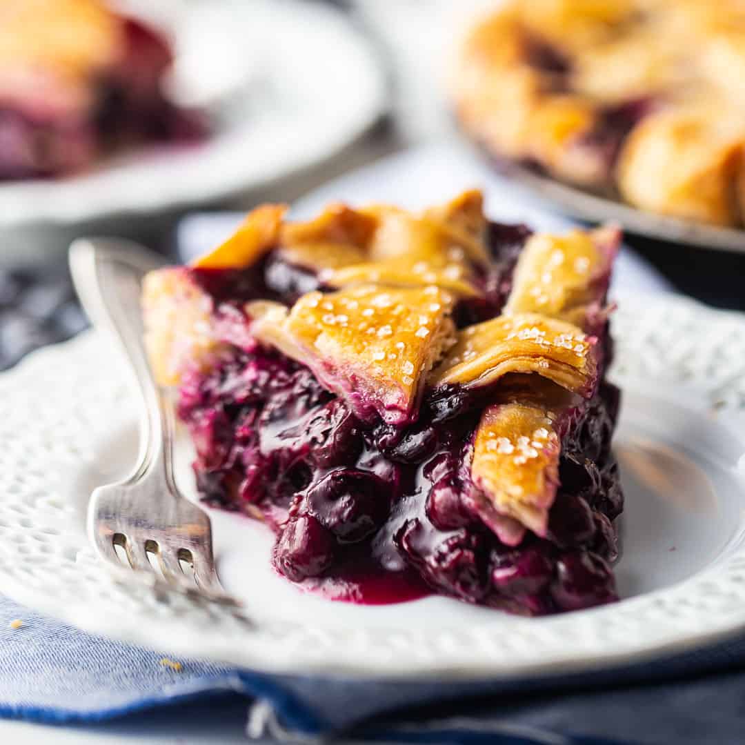 A slice of blueberry pie with thick filling and a lattice top crust, presented on a lacy white plate.