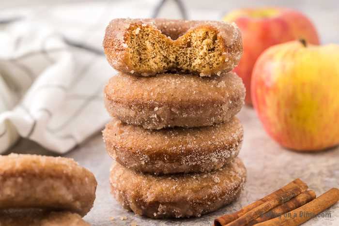 Apple Cider donuts stacked on top of each other with a bite taken out of the top donut with apples behind it.