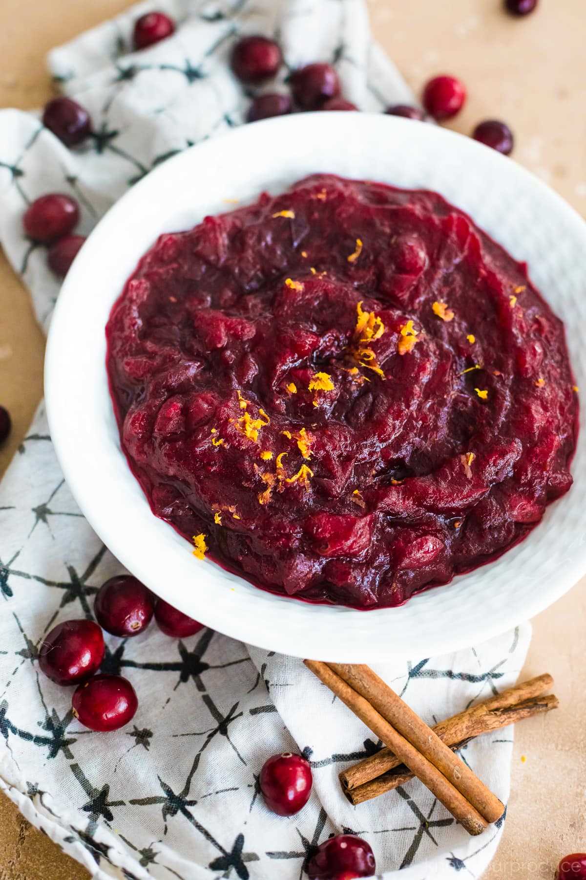 overhead fresh cranberry sauce in a white bowl with a napkin under it and cranberries and cinnamon sticks scattered