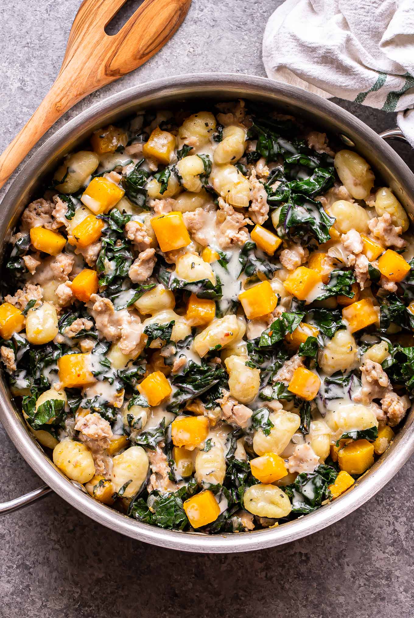 Butternut Squash, Sausage, and Kale Gnocchi in a stainless steel Skillet with a wooden serving spoon behind it.