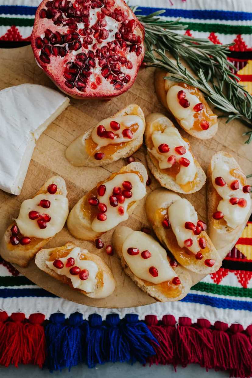 9 pieces of brie bruschetta crostini on a round wooden cutting board with 2/3 a wedge of brie, a halved pomegranate and fresh sprigs of rosemary.